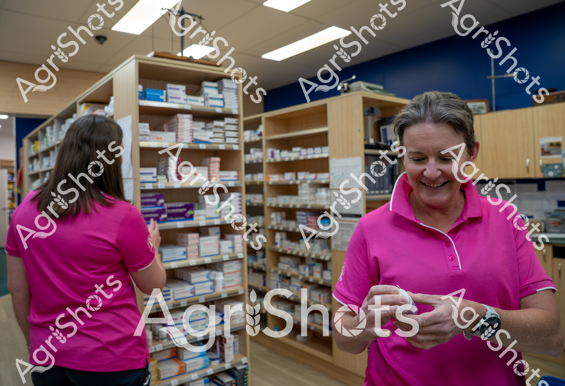 Two pharmacy workers in bright pink shirts work among shelves stocked with numerous medication boxes and prescription medicines. One woman faces away while the other smiles and handles a medication container in what appears to be a well-organized pharmacy storage or inventory area with wooden shelving and blue walls.