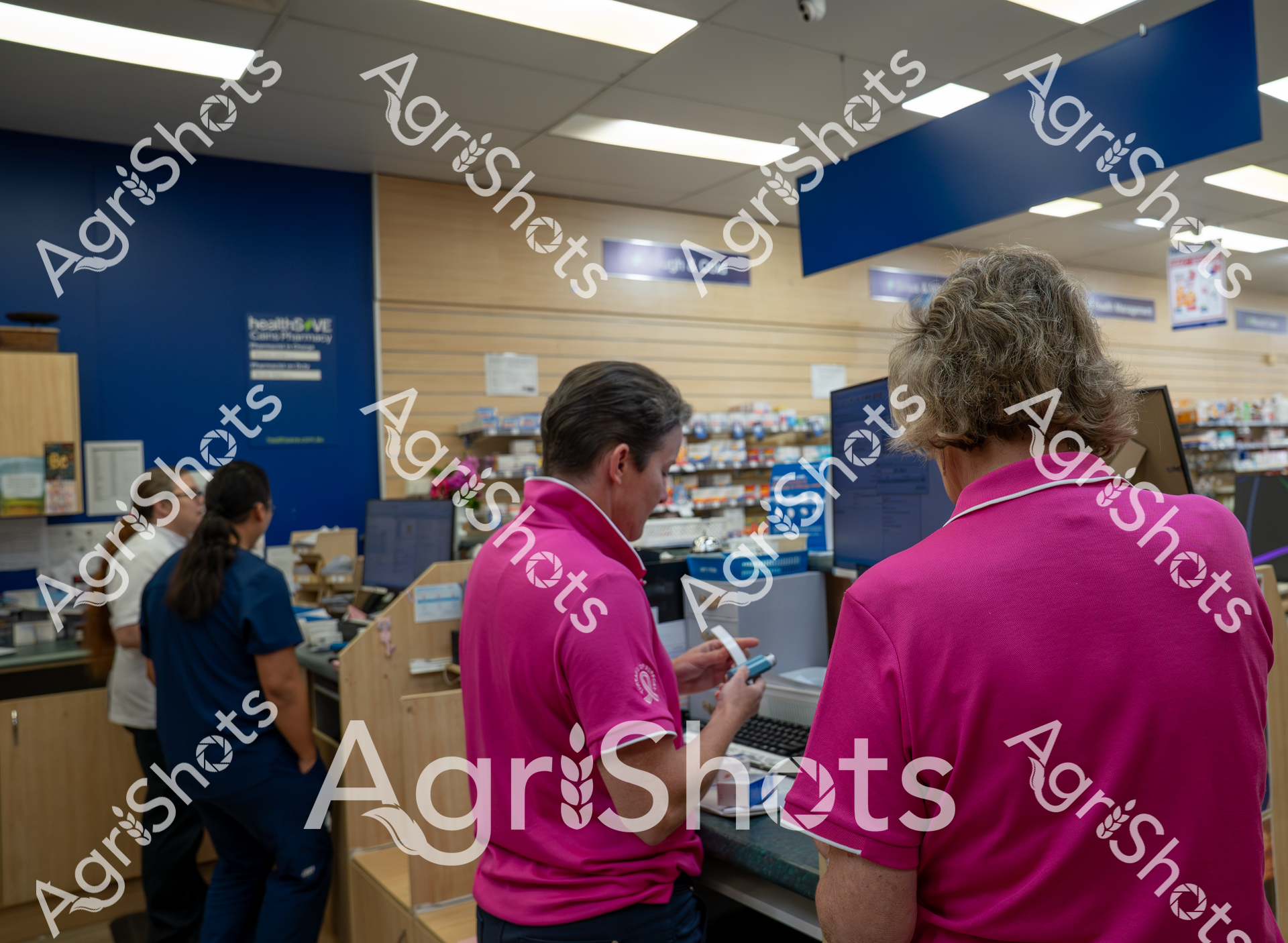 Two pharmacy staff in bright pink shirts work at a computer terminal in a pharmacy setting, with blue walls and shelves of medication visible in the background. They appear to be processing prescriptions or assisting a customer at the counter.