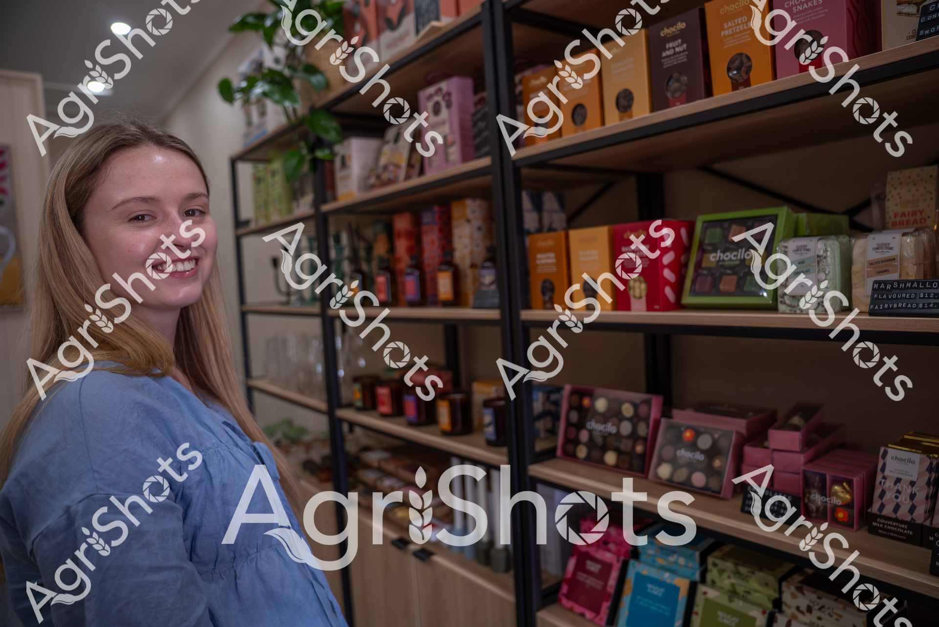 A smiling young woman in a blue top stands in a specialty store, surrounded by shelves filled with colorful chocolate and confectionery products. Her warm expression and the inviting display of artisan treats suggest a passion for gourmet sweets in this cozy retail environment.