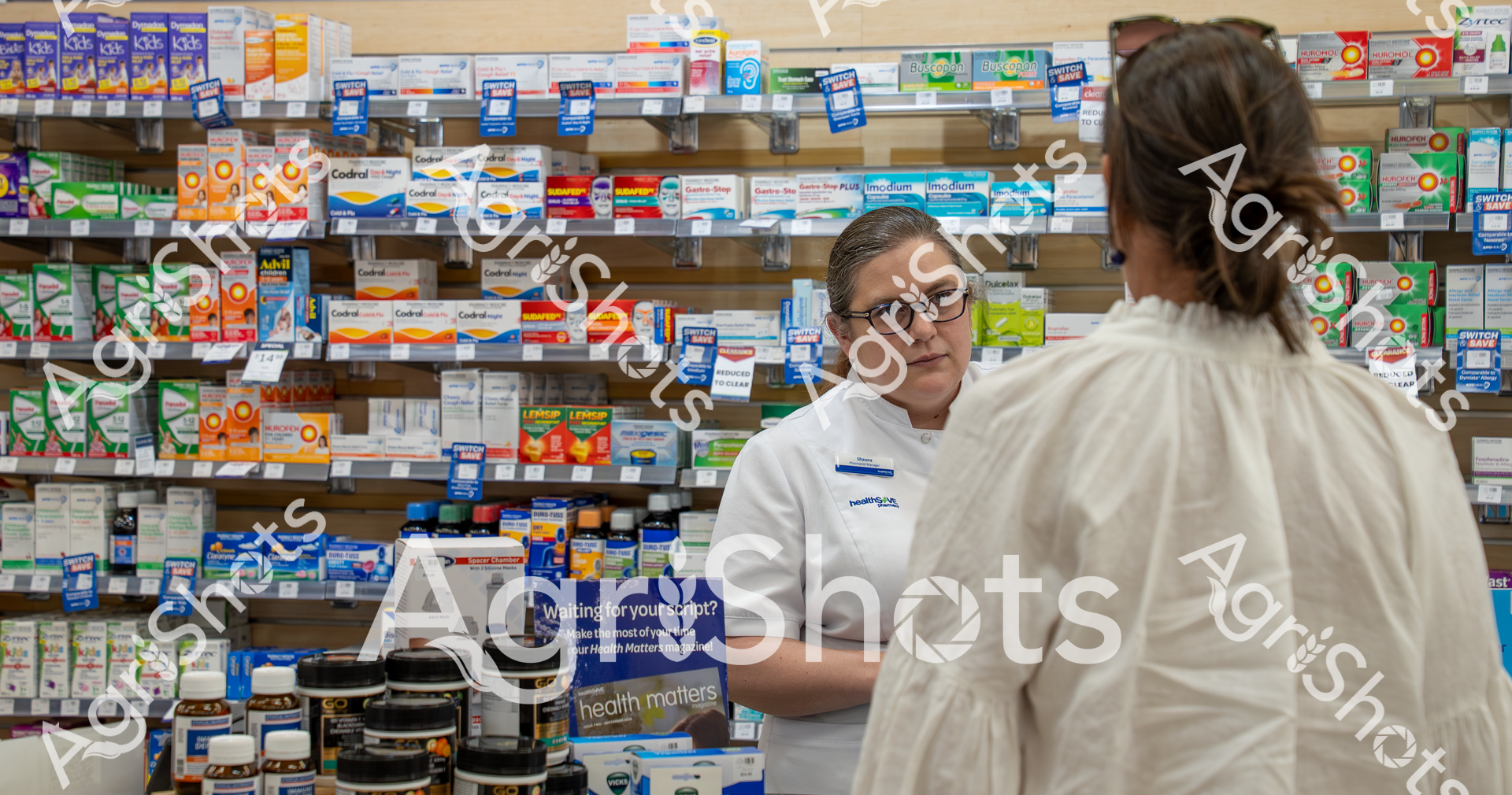 A pharmacist in a white coat stands behind a counter filled with numerous medication boxes and bottles, helping a customer. The pharmacy shelves are densely packed with various over-the-counter and prescription medicines, showcasing the wide range of health products available.