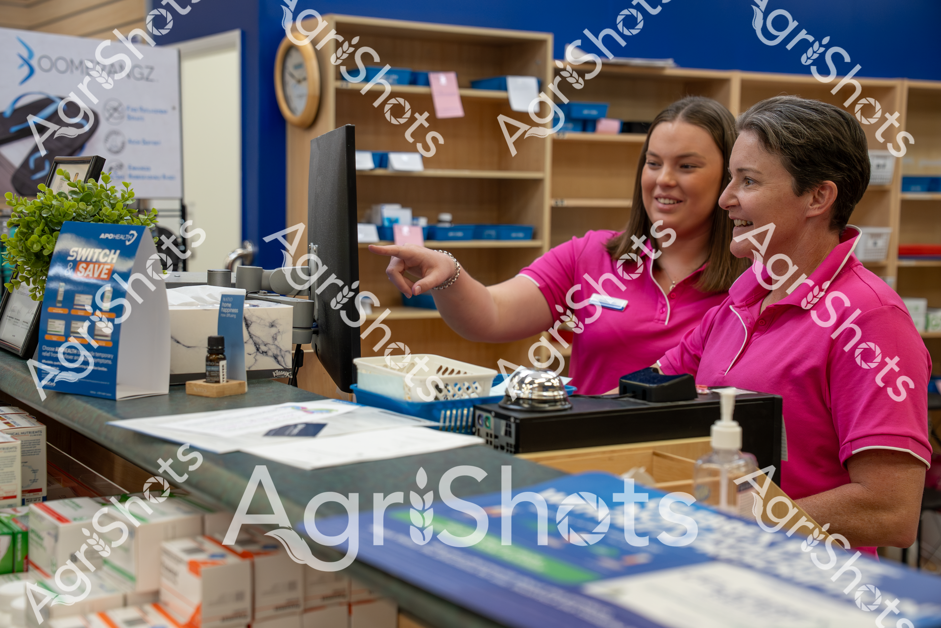 Two women in matching bright pink shirts work together at a retail counter, smiling and appearing to collaborate on a task. They are in a store with shelves and product displays visible, and seem engaged in friendly conversation while working.