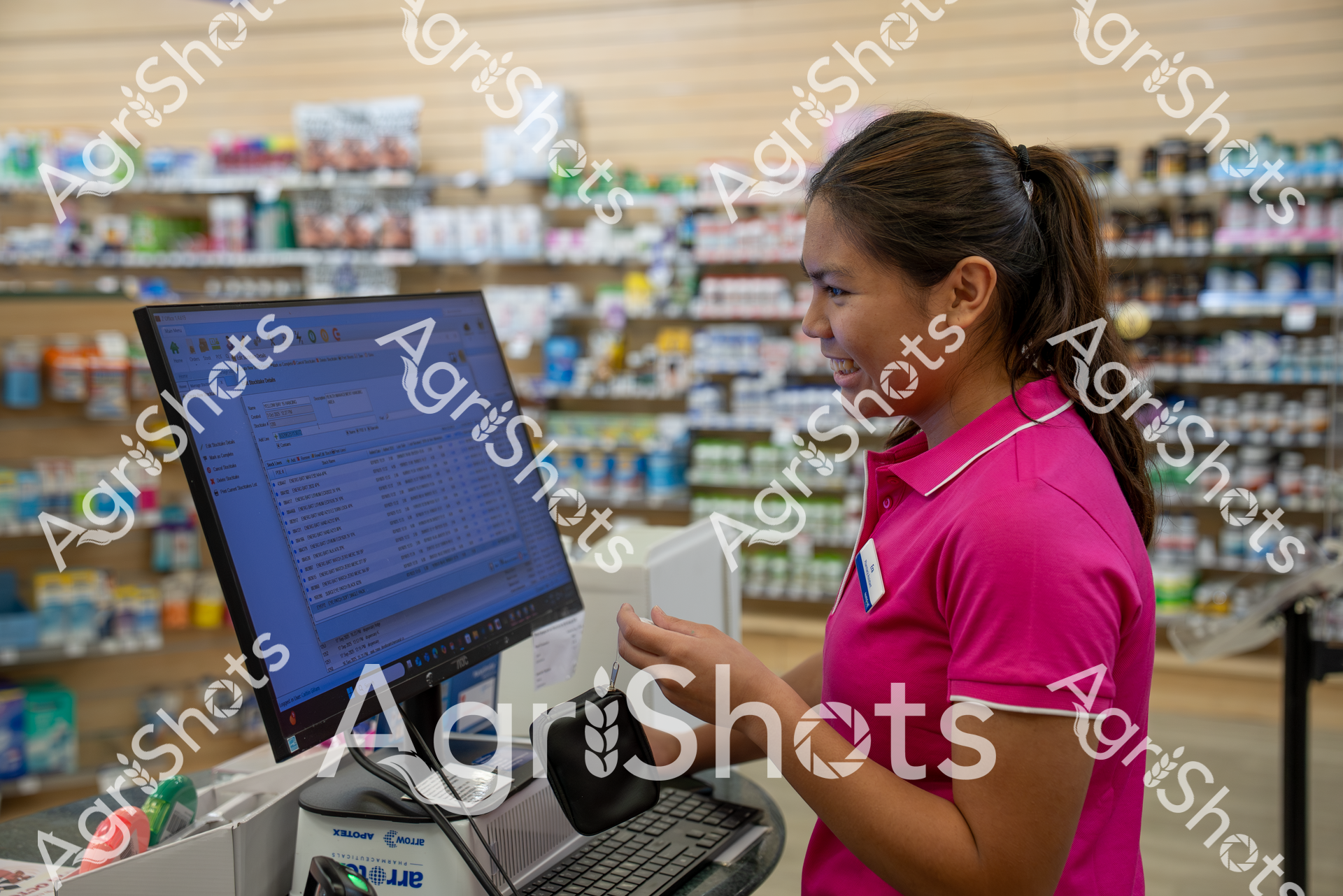 A woman in a bright pink shirt works at a pharmacy computer, surrounded by shelves of medical products and medications. She appears focused on the screen, likely processing prescriptions or checking inventory in the store.
