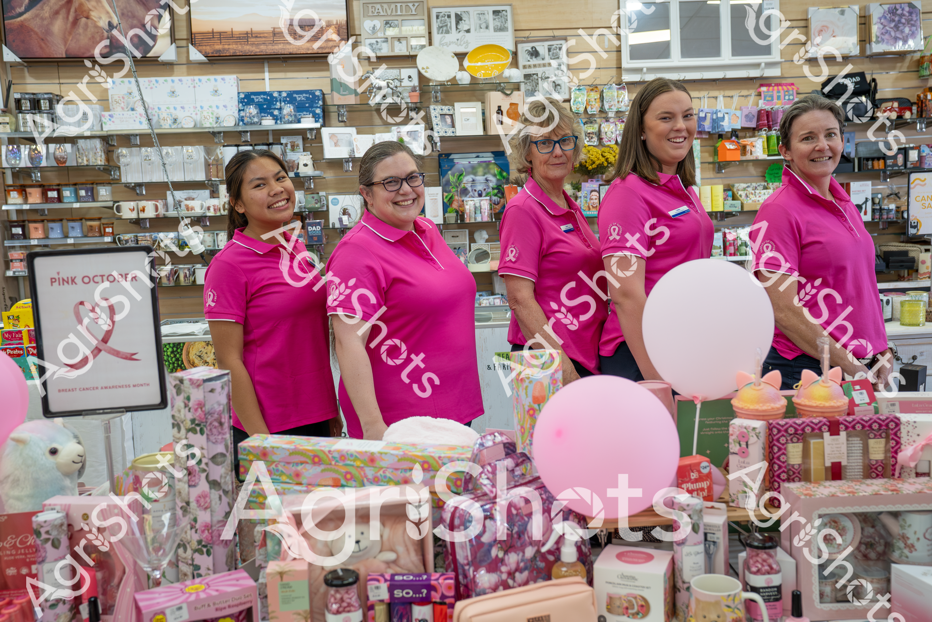 Five women wearing bright pink polo shirts stand together in a gift shop, surrounded by pink balloons and merchandise. They are smiling and appear to be participating in a breast cancer awareness event or campaign. The store's background is filled with various gift items, frames, and decorative products.