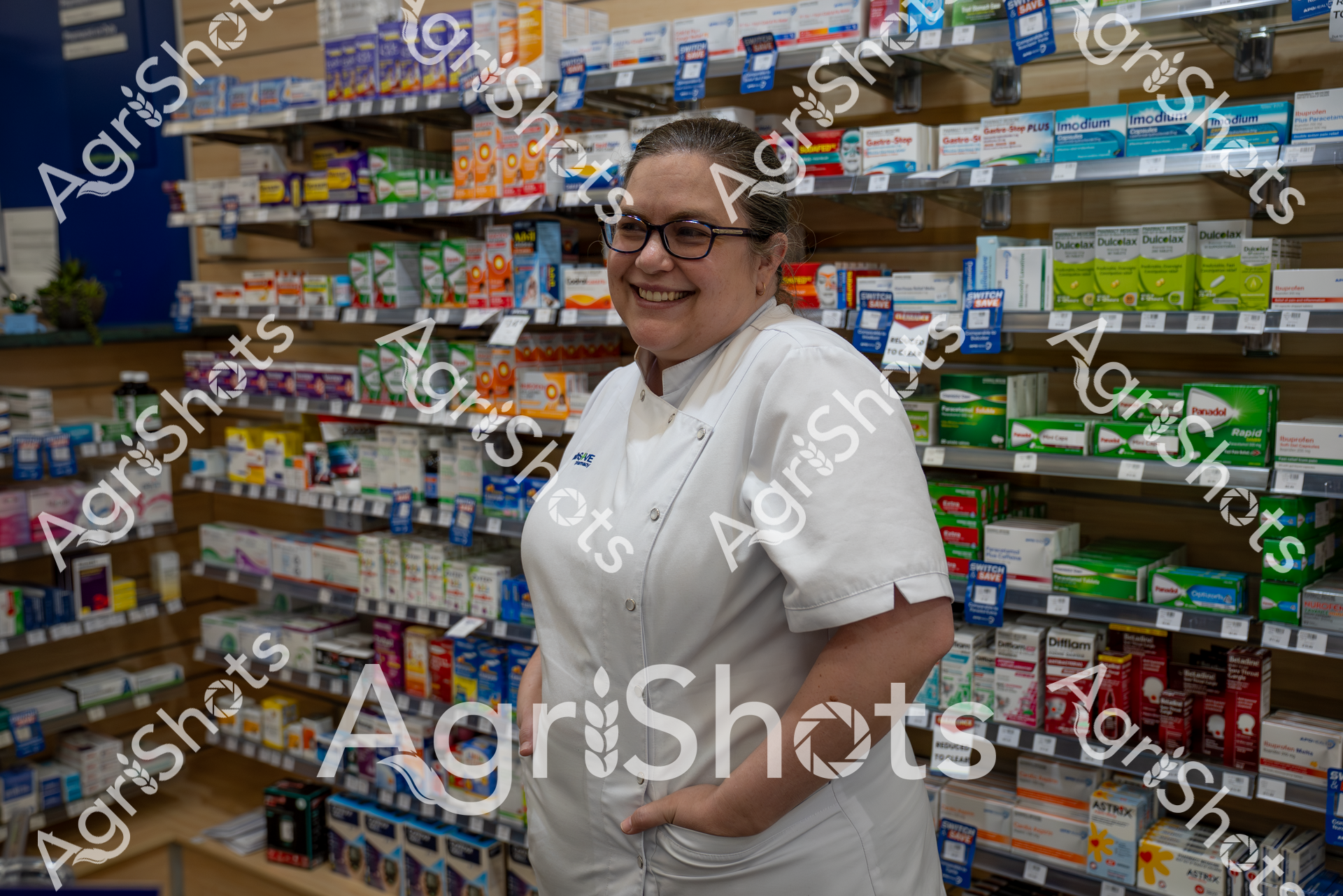 A smiling woman in a white pharmacy uniform stands in a pharmacy, surrounded by shelves filled with various medications and healthcare products. She appears confident and welcoming, wearing glasses and with her hair pulled back, embodying a professional and caring demeanor in this healthcare setting.