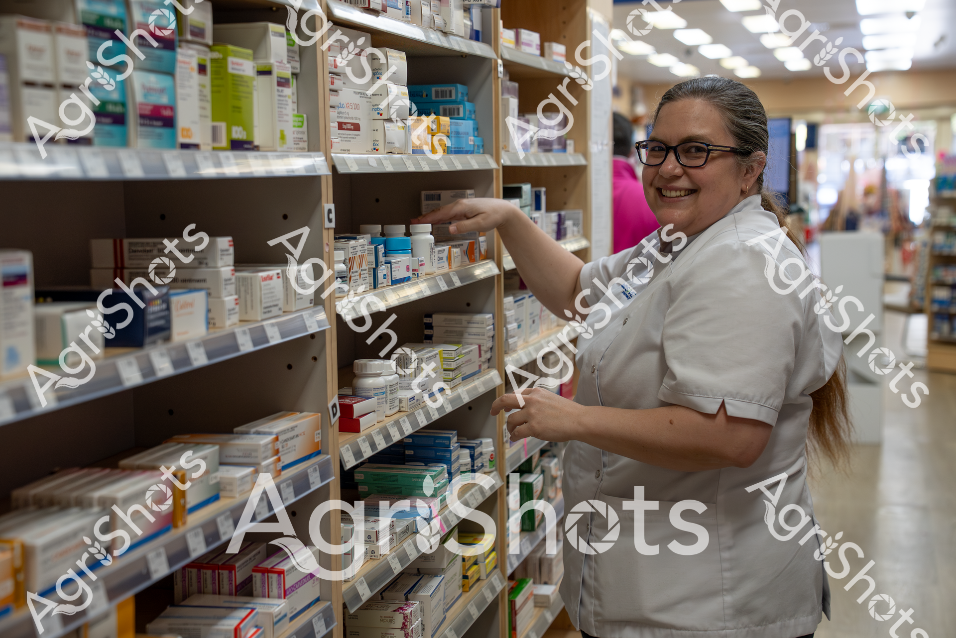 A smiling woman in a white uniform and glasses stands in a pharmacy, reaching for medication on shelves filled with various pharmaceutical products. She appears to be a pharmacist or pharmacy technician organizing and managing the inventory of prescription drugs and medical supplies.