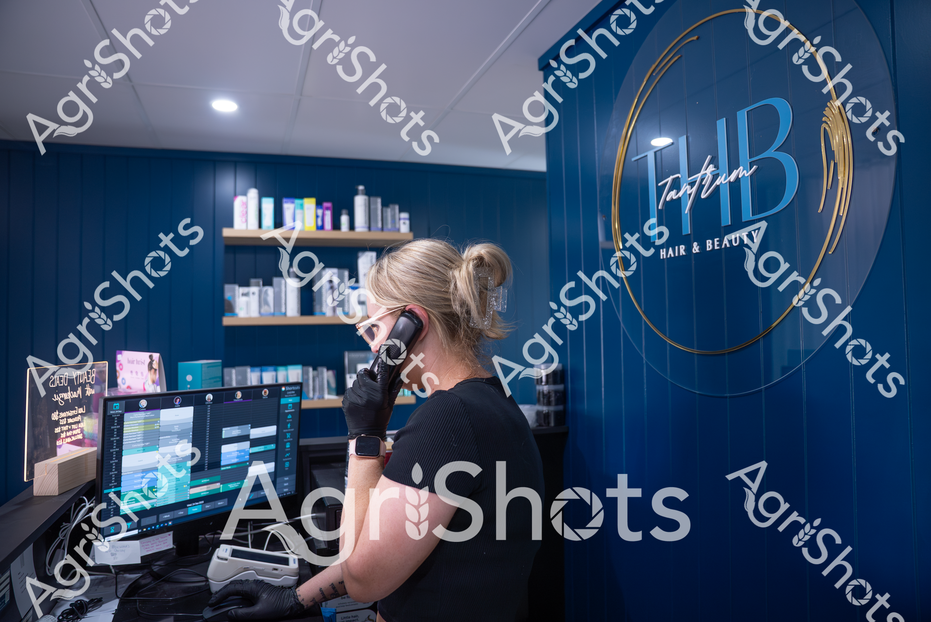 A woman with blonde hair pulled back works at a computer in a modern hair and beauty salon. She is wearing black and using a phone while sitting at a workstation with multiple screens. The salon, called HB Hair & Beauty, has a sleek blue interior with product shelves in the background.