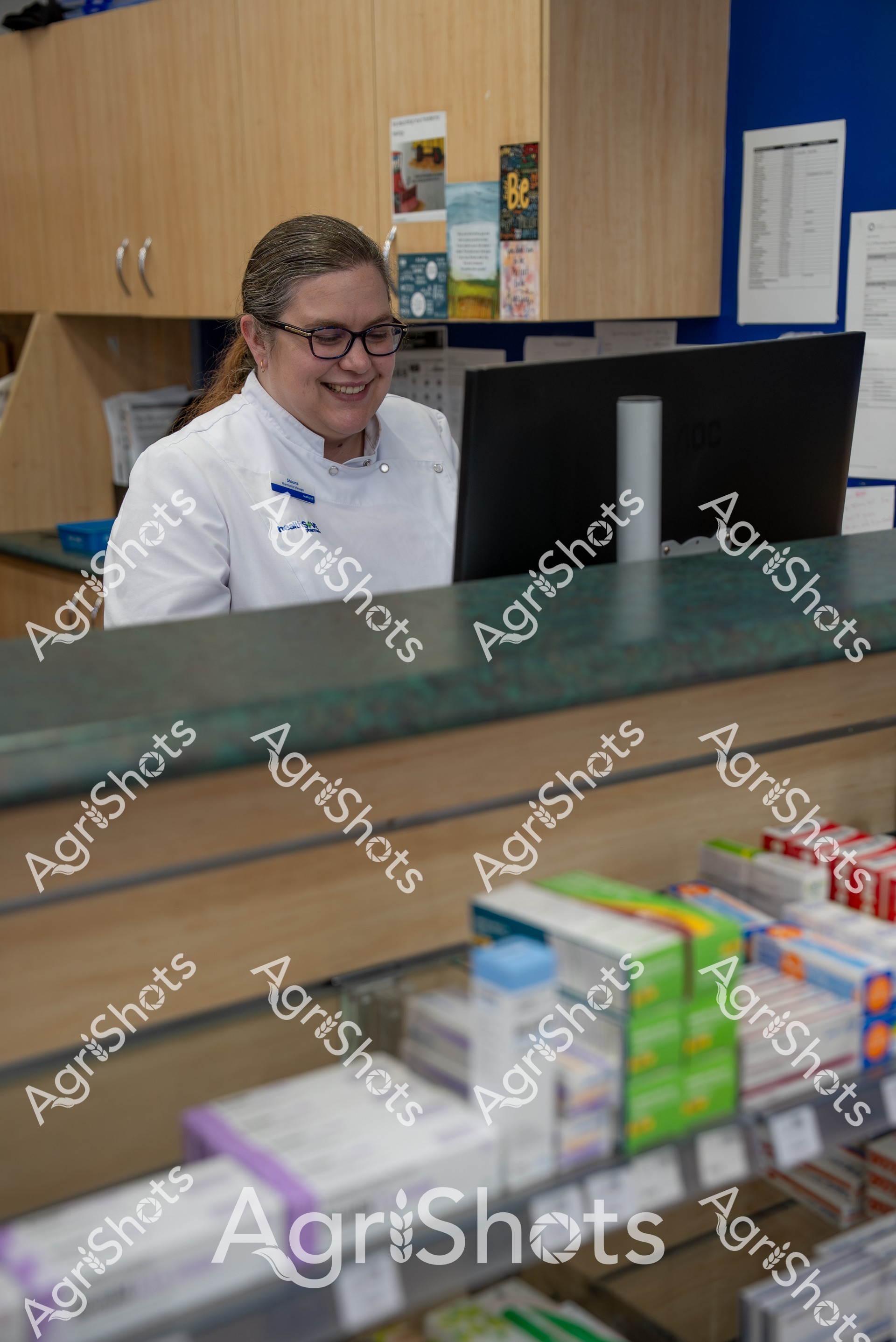 A woman wearing glasses and a white professional coat smiles while working at a computer in what appears to be a pharmacy or medical setting. Several shelves of medication boxes are visible in the foreground, and wooden cabinets and bulletin boards can be seen in the background, suggesting a healthcare workplace environment.