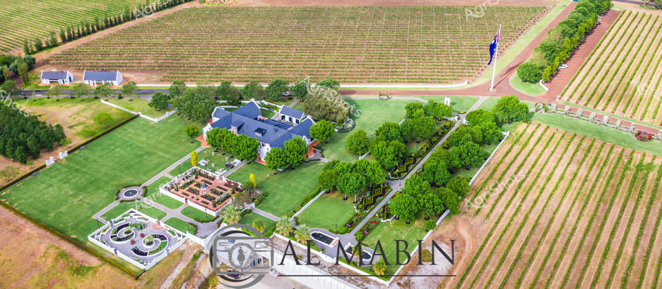 Aerial panorama of an immaculate Cape Dutch-style winery estate nestled amid golden wheat and verdant grape rows in Margaret River's prestigious wine region. Meticulously landscaped gardens radiate geometric precision, with manicured lawns, symmetrical tree plantings, and an elegant circular water feature framing the slate-roofed homestead. Neat vineyard blocks stretch to the horizon, revealing the careful agricultural planning of this premium wine-producing landscape. The property exemplifies Western Australian viticultural excellence, with precisely aligned vine rows contrasting against carefully maintained green spaces and architectural elegance. Soft morning light bathes the scene, highlighting the rich terra cotta soil, emerald green lawns, and the orderly rows of agricultural productivity that define this remarkable wine-growing terrain. The image captures the harmonious intersection of agricultural science, architectural design, and natural beauty that characterizes Margaret River's world-renowned wine estates.