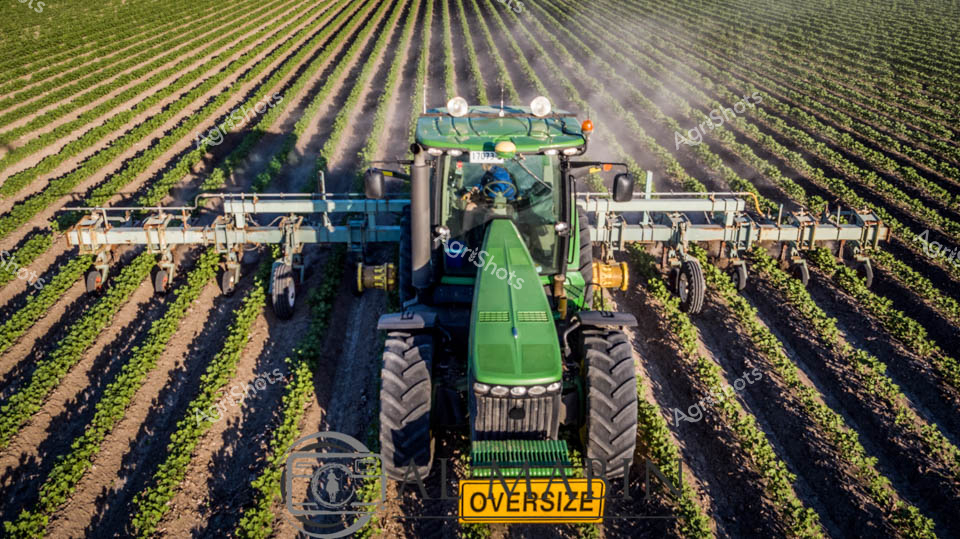 Precision agriculture unfolds across a meticulously planted cotton field, with a gleaming green John Deere tractor navigating perfectly aligned rows of young crops. The expansive agricultural landscape stretches into the distance, revealing mathematically straight planting lines that demonstrate cutting-edge farming techniques. Mounted with an extensive multi-row cultivation implement, the oversized tractor straddles the delicate green seedlings, its bright yellow 'OVERSIZE' warning sign highlighting the specialized machinery's impressive scale. Early morning light catches the dust and mist rising around the vehicle, creating a soft atmospheric haze that emphasizes the rhythmic geometry of the plantation. The tractor's advanced suspension and precision GPS guidance system allows for minimal soil disruption, representing the pinnacle of modern agricultural technology and sustainable farming practices. Every element speaks to Queensland's sophisticated agricultural sector - where technology, skill, and environmental awareness converge to nurture future crop yields.