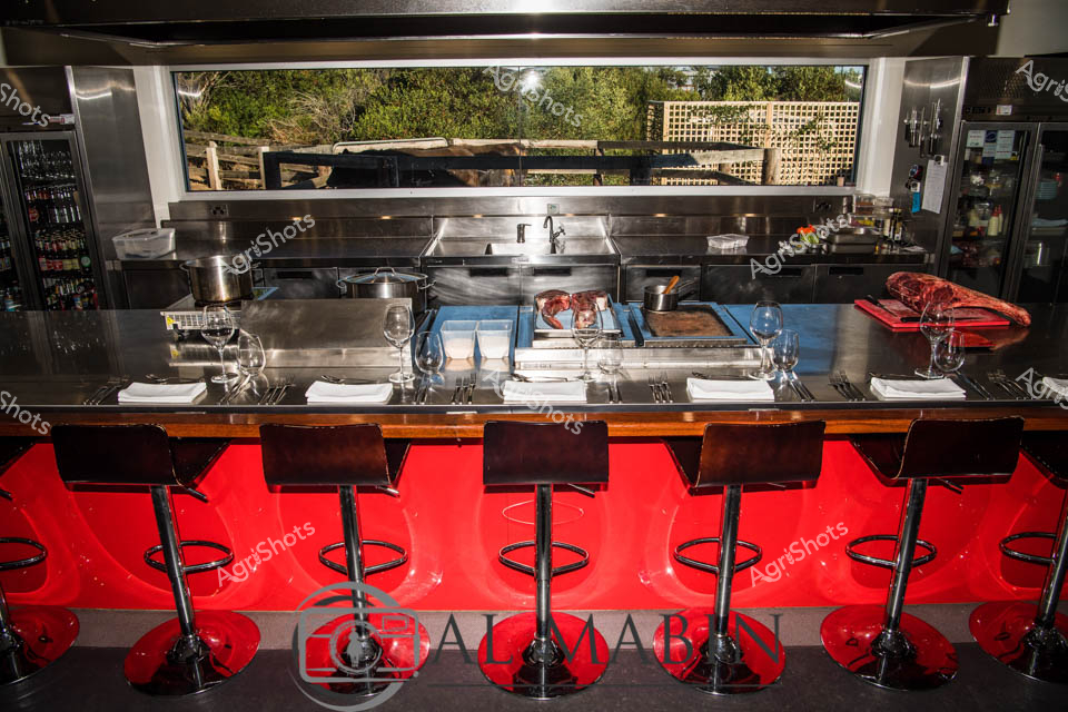 Gleaming stainless steel and vibrant red create a theatrical stage for culinary performance at this high-end restaurant kitchen bar. Premium Wagyu beef cuts rest prominently on a pristine preparation surface, showcasing the pinnacle of South Australian agricultural excellence. Precisely arranged wine glasses and crisp white linens frame an immaculate workspace that blends professional kitchen functionality with elegant dining design. Sophisticated bar stools in deep burgundy and black invite diners to witness the artistry of meat preparation, with a glimpse of lush greenery visible through the expansive kitchen window. The meticulously organized space speaks to the precision of Australian agricultural production, from pasture to plate. Every element - from the expertly displayed beef to the carefully positioned culinary tools - demonstrates a commitment to quality that defines South Australia's premium food culture.
