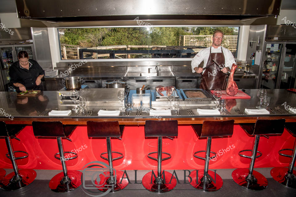 Open kitchen theatre showcases the precision of premium beef preparation at a high-end restaurant. A professional chef in a crisp white uniform and burgundy leather apron carefully handles a large primal cut of deep red Wagyu beef, highlighting the premium quality of South Australian cattle production. Gleaming stainless steel surfaces and professional cooking equipment create a clinical, artistic workspace, with red bar stools adding a vibrant accent to the professional kitchen environment. Another kitchen staff member works quietly in the background, preparing complementary ingredients with focused concentration. Elegant wine glasses and pristine white linens suggest a sophisticated dining experience that celebrates the finest local beef production. The expansive kitchen window reveals a glimpse of natural landscape, connecting the refined culinary preparation to the agricultural origins of the premium meat.