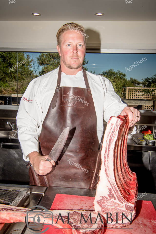 A professional chef stands confidently in a modern kitchen, wearing a crisp white chef's jacket and a rich brown leather apron. He holds a traditional butcher's knife in one hand and grips a premium cut of marbled Wagyu beef in the other, revealing the exceptional quality of the meat with its distinctive red and white intramuscular fat patterns. The scene speaks to the precision of high-end culinary preparation, showcasing the journey from premium livestock to gourmet dining. Recessed lighting and glass windows behind him create a clean, professional backdrop that highlights the deep red tones of the beef and the chef's focused demeanor. Every element of the composition communicates excellence in meat preparation - from the chef's expert grip to the pristine cutting surface, embodying the artistry of modern Australian agricultural and culinary expertise.