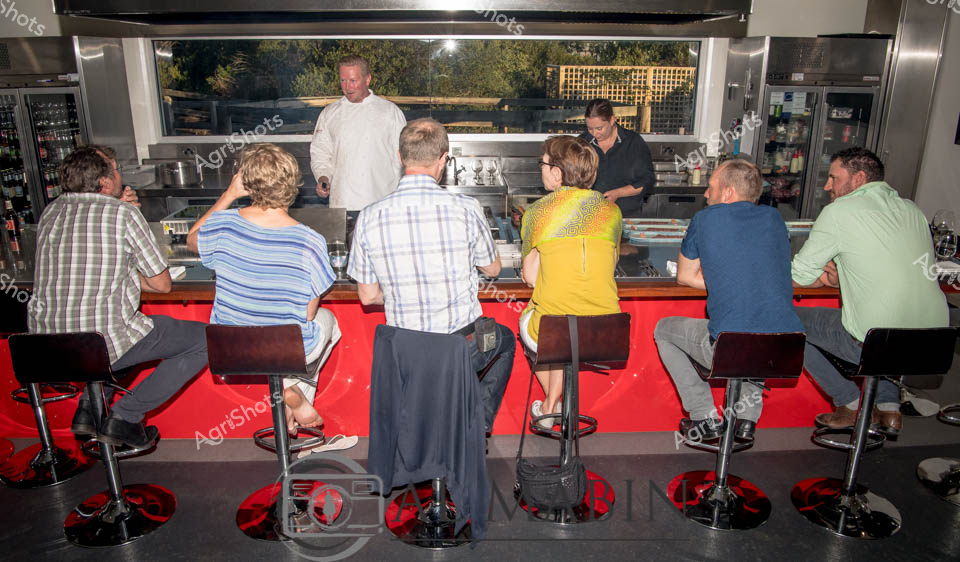 Professional kitchen chefs and staff gather at a sleek counter, observing an expert chef in white preparing a culinary demonstration. The kitchen features a vibrant red counter with black bar stools, creating a dynamic staging area for a cooking class or behind-the-scenes restaurant experience. Stainless steel equipment and glass-fronted refrigerators line the background, with natural light filtering through a window revealing an outdoor latticed area. The participants - dressed in casual shirts ranging from plaid to striped to solid colors - are seated intently, suggesting a professional training or specialized food preparation workshop. The scene captures the precision and collaborative spirit of high-end culinary preparation, with an atmosphere of focused learning and gastronomic expertise. Gleaming surfaces, professional kitchen attire, and the attentive postures of the participants convey a sense of serious culinary craftsmanship and skill development.