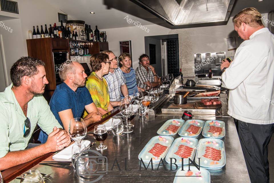 Precision butchery and culinary expertise converge in this intimate restaurant kitchen scene, showcasing premium Wagyu beef preparation. A skilled chef in crisp white attire stands at a professional-grade cooking station, meticulously arranging vibrant red meat portions across blue serving platters. A group of engaged patrons lines the sleek kitchen bench, wine glasses in hand, observing the intricate process of meat selection and preparation with rapt attention.

The refined interior blends industrial and contemporary design, with a well-stocked bar of wine bottles visible in the background and a reflective metallic ceiling creating an atmosphere of sophisticated gastronomy. Soft lighting and the careful arrangement of ingredients suggest an exclusive dining experience focused on high-quality beef - likely sourced from renowned South Australian Wagyu producers like Mayura Station.

Each participant appears deeply invested in the culinary demonstration, representing a confluence of agricultural expertise and fine dining. The color palette - from the chef's pristine white uniform to the subtle earth tones of the patrons' clothing and the rich red of the meticulously prepared beef - creates a visual harmony that speaks to the precision of Australian agricultural and culinary craftsmanship.