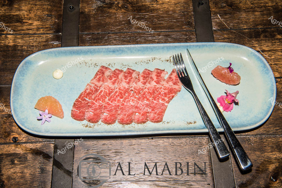 Marbled ruby-red Wagyu beef slices cascade across a pale blue ceramic platter, showcasing the exquisite marbling characteristic of premium South Australian cattle breeding. Delicate pink and orange flower petals and citrus segments artfully accent the meat, suggesting a refined culinary presentation that celebrates the region's high-end agricultural produce. The precision-cut beef gleams with intricate fat striations, representing the pinnacle of Mayura Station's renowned Wagyu genetics and meticulous husbandry. Elegant black cutlery frames the composition, hinting at a sophisticated dining experience that transforms agricultural excellence into gastronomic artistry. This image captures the seamless journey from pasture to plate, highlighting the state's world-class beef production and the intricate craft of transforming livestock into gourmet cuisine.