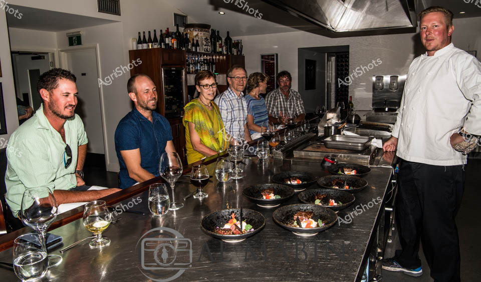 Fine dining meets agricultural precision in this intimate culinary workshop. A professional chef in crisp white attire stands alongside a small group of guests at a sleek stainless steel kitchen bench, with carefully plated gourmet dishes suggesting a high-end tasting experience. The diverse group of diners - dressed in casual business and weekend attire - lean in with wine glasses, suggesting a curated gastronomic event that celebrates sophisticated food preparation. Dark-toned ceramic plates feature intricate dishes that hint at premium agricultural produce, likely showcasing premium South Australian beef, possibly from renowned Wagyu or Mayura Station cattle. The sophisticated bar backdrop, with an extensive wine collection, and the professional kitchen setup create an atmosphere of refined agricultural storytelling, where food becomes an art form connecting producers and consumers. Soft lighting and the attentive postures of participants underscore the precision and passion behind this culinary experience.
