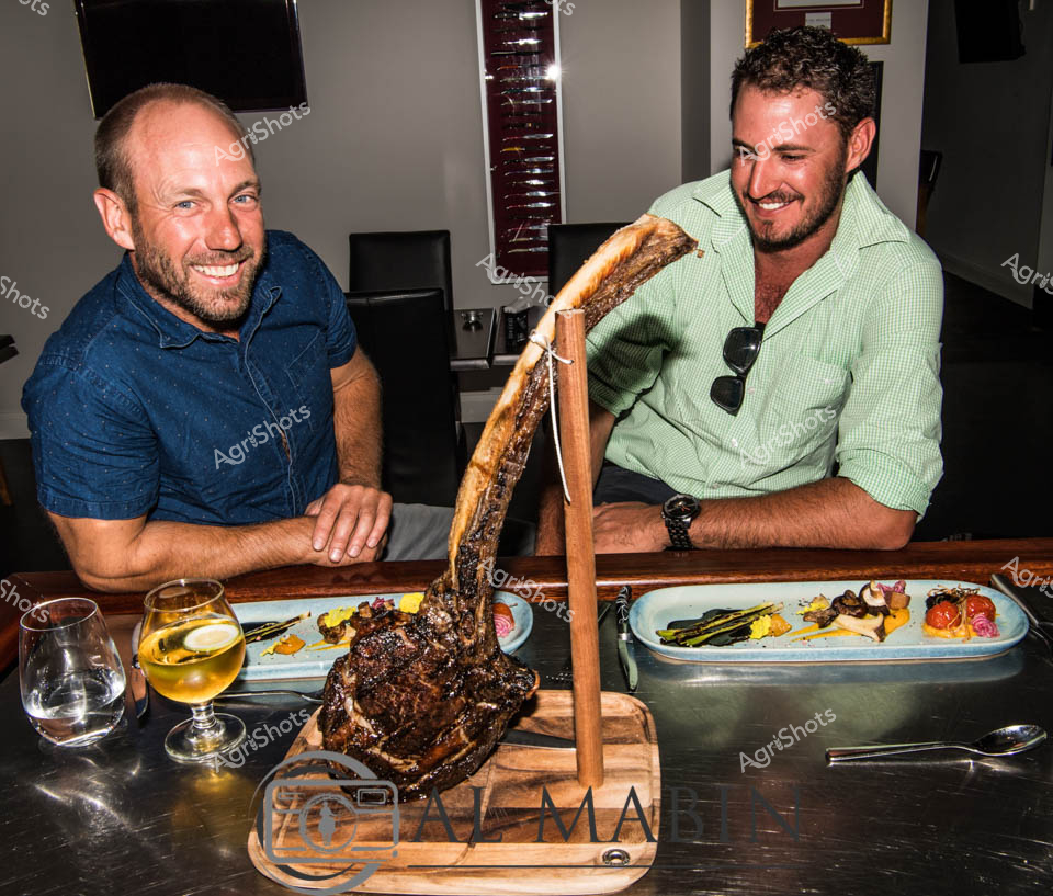 Succulent Wagyu beef takes center stage in this intimate culinary moment, showcasing the pinnacle of Australian beef production. A massive, perfectly seared tomahawk steak hangs dramatically from a wooden presentation stand, suspended between two enthusiastic diners in a refined restaurant setting. The men - one in a crisp blue polo, the other in a light green gingham shirt - radiate excitement and appreciation for this premium cut, likely sourced from Mayura Station's renowned Wagyu feedlot in South Australia. Accompanying the centerpiece are carefully plated side dishes featuring vibrant yellow and green vegetables, and glasses of white wine that catch the light. Their expressions reveal the pride and joy of Australia's premium beef industry, bridging the journey from carefully raised cattle to an exquisite dining experience that celebrates agricultural craftsmanship. The wooden serving board and carefully curated plating highlight the premium quality of this locally produced beef, embodying the sophisticated farm-to-table ethos of modern Australian cuisine.