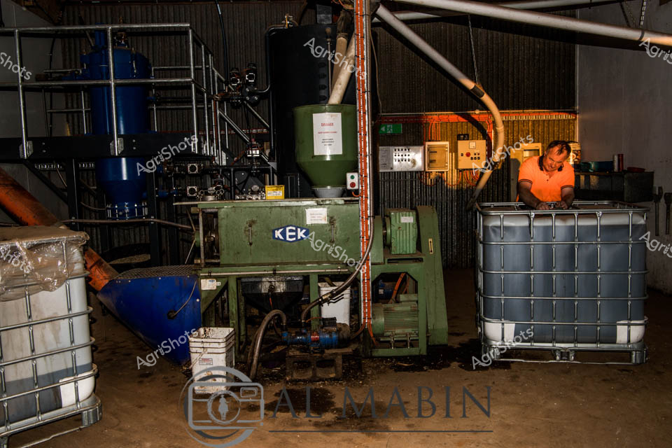 A worker in an orange safety shirt operates specialized poultry processing equipment inside an industrial farm facility in Tasmania. The green KEK machinery, surrounded by large storage containers and processing tanks, forms part of an large-scale egg production operation. Blue storage vessels and white industrial containers are positioned throughout the workspace, highlighting the scale of modern commercial poultry farming. The indoor facility showcases the mechanized infrastructure required for large-scale chicken and egg production systems.