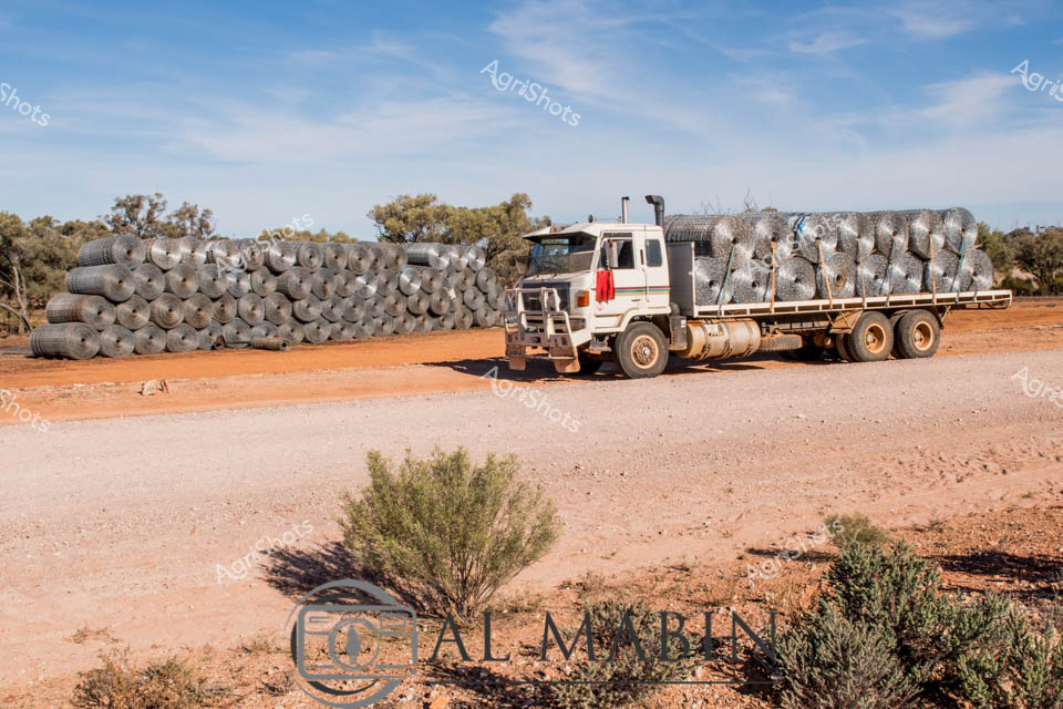 A robust, white Mack truck sits against the rich, rusty-red earth of the Queensland outback, loaded with neatly bundled rolls of wire mesh fencing. Scattered saltbush and hardy scrub punctuate the arid landscape, under a soft blue sky with wispy cirrus clouds suggesting a mild late winter or early spring morning. The truck's flatbed is stacked high with uniform cylindrical rolls of galvanized wire mesh, essential for maintaining boundary fences across vast pastoral stations. This scene captures the logistical backbone of rural Australian agriculture - the critical task of fence maintenance that preserves property boundaries and manages livestock across immense, challenging terrains. The steel-gray wire rolls contrast beautifully with the ochre ground, symbolizing human resilience and technological adaptation to harsh environmental conditions. Every element speaks to the methodical, strategic approach required to manage agricultural operations in remote Australian landscapes, where infrastructure and preparedness are key to survival.