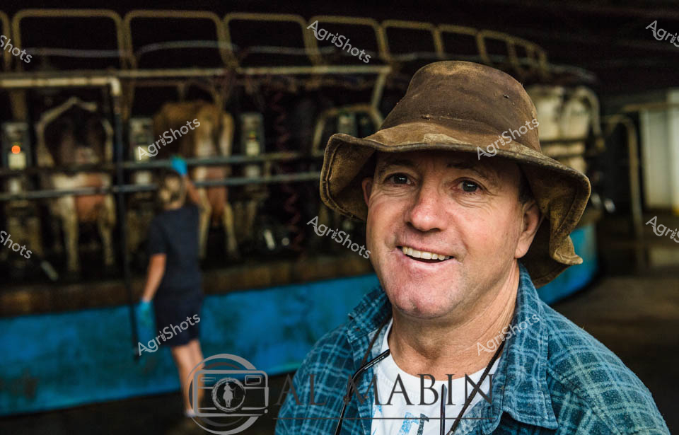A smiling man working as a dairy farmer stands in front of a rotary milking parlor in Western Australia, wearing a weathered hat and blue checkered shirt. The circular milking system is visible in the background with its characteristic metal framework and blue equipment panels. His cheerful expression reflects satisfaction with the modern dairy operation, showcasing the efficient rotary dairy technology used for milking dairy cows in this agricultural setting.