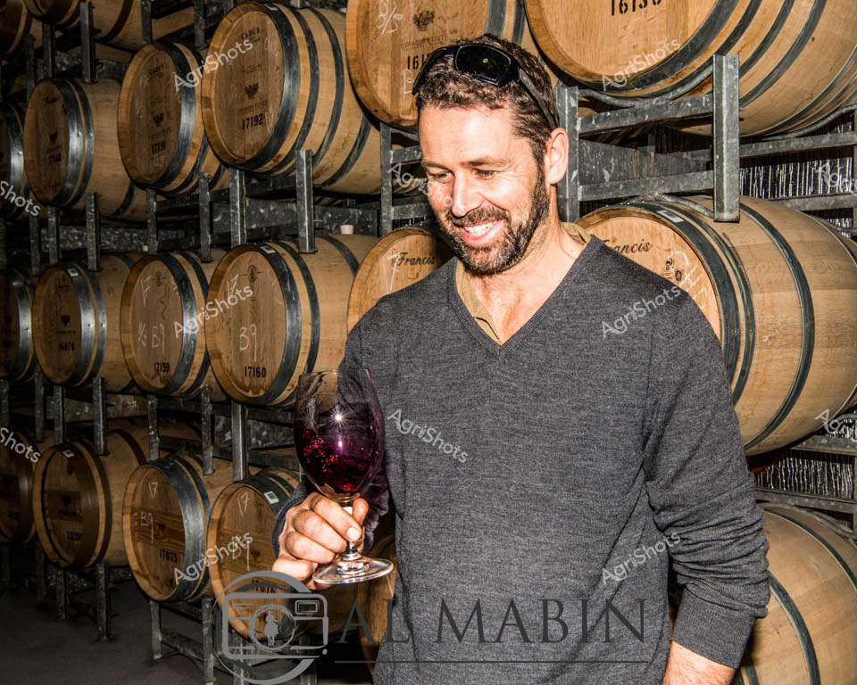 A passionate vintner stands amid towering oak wine barrels in a classic barrel room, holding a glass of deep crimson red wine. Soft ambient light illuminates the rich wooden textures of carefully stacked wine barrels, each marked with unique identification numbers and winery codes. The individual wears a comfortable grey V-neck sweater, his relaxed posture and warm smile reflecting the refined yet approachable culture of Margaret River's renowned wine region in Western Australia. Rows of meticulously arranged French oak barrels create a rhythmic backdrop, suggesting the patient craft of wine aging and the sophisticated agricultural heritage of the region. The deep burgundy wine swirls gently in the glass, catching the light and hinting at the complex flavors developing within the wooden vessels surrounding him. This scene captures the essence of Western Australian viticulture - a blend of technical precision, artistic passion, and deep respect for the natural processes that transform humble grapes into world-class wines.