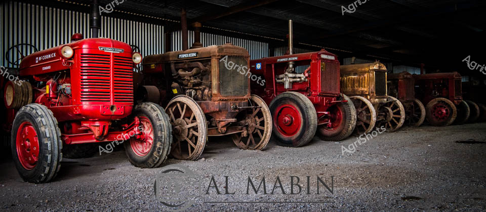 Scarlet and rust-toned vintage tractors line the corrugated iron shed, a testament to agricultural heritage preserved with meticulous care. The McCormick tractors, ranging from vibrant red to weathered bronze, stand as stoic sentinels of farming history, their robust wheels and intricate mechanical details telling stories of decades of rural labor. Soft industrial lighting cascades across the machinery, highlighting the textured patina of aged metal and worn paintwork that speaks to years of challenging field service. Each tractor represents a different era of agricultural innovation, carefully arranged to showcase the evolution of farming technology from the mid-20th century. The gravel floor and metallic wall backdrop create a timeless museum-like atmosphere, transforming these working machines into revered artifacts of Australia's agricultural legacy. The signature at the bottom of the image signals this as a carefully curated photographic celebration of rural mechanical engineering.