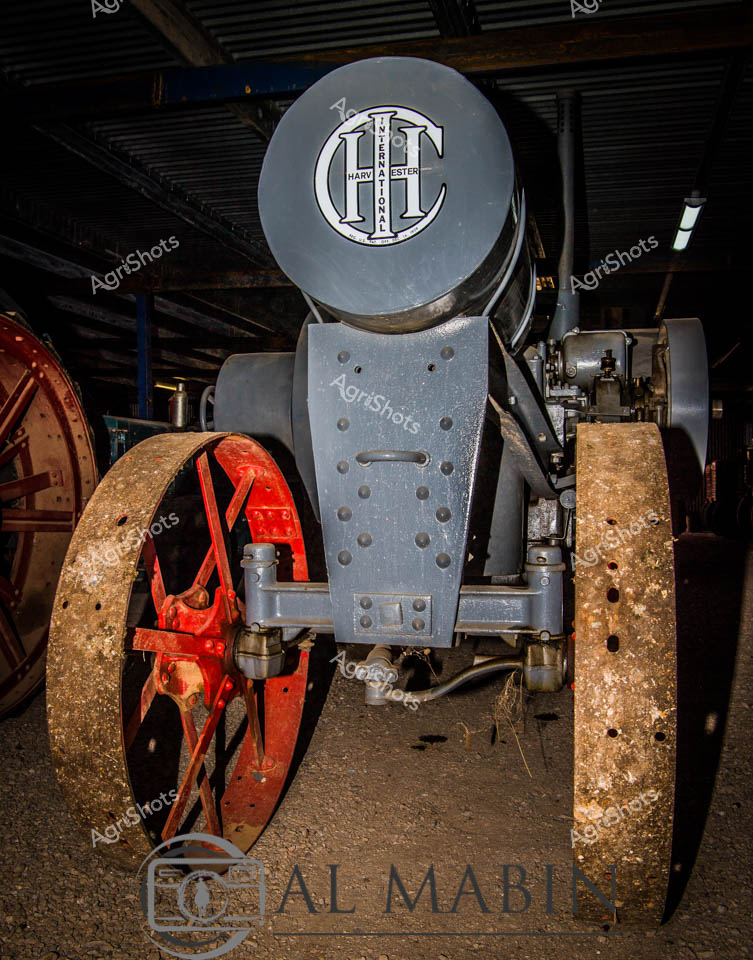 Immortalized in the dimly lit sanctuary of an agricultural heritage museum, this International Harvester tractor stands as a gleaming testament to early 20th-century industrial engineering. The machine's robust frame is anchored by massive golden-rusted wheels, their weathered surfaces telling stories of countless paddocks traversed, contrasting dramatically with the brilliant red wheel hubs that pulse with mechanical vitality. Riveted steel panels in steely grey form the tractor's skeletal structure, punctuated by careful rows of bolts that speak to the precision engineering of its era. The prominent International Harvester logo crowns the machine, a proud emblem of agricultural innovation that transformed farming across rural Victoria and beyond. Shadows and ambient museum lighting caress the mechanical contours, highlighting every mechanical nuance and casting an almost reverential glow on this magnificent piece of agricultural machinery. This vintage tractor represents more than mere equipment - it embodies the technological spirit and mechanized revolution that reshaped Australian agricultural practices in the early decades of the 20th century.