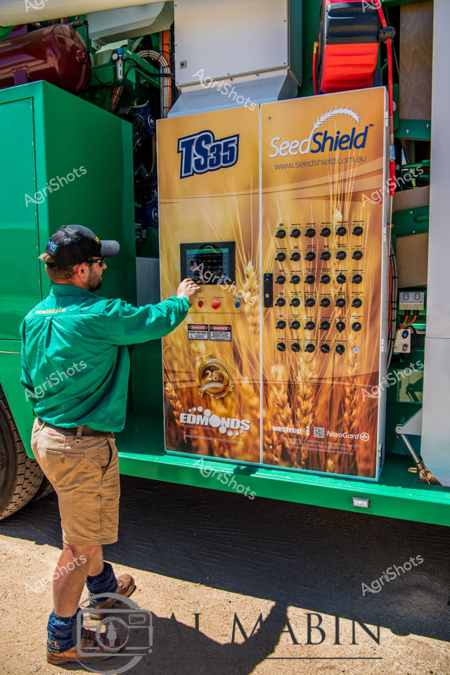 A man working as a farm equipment operator adjusts controls on the bright yellow SeedShield TS35 control panel mounted on green agricultural machinery. The technician wears a green work shirt while operating the seed cleaning equipment, with its array of switches and digital displays clearly visible. The scene captures modern cropping technology in Western Australia's agricultural sector, showcasing the precision equipment used for seed processing and harvest operations.