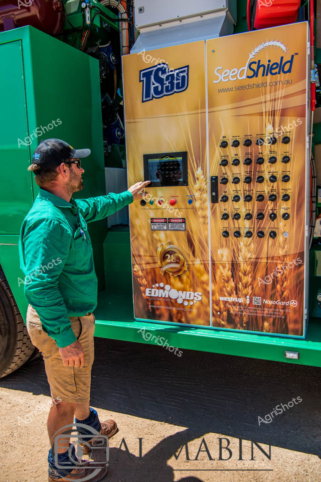 A man working as a farm operator interacts with the control panel of a TS95 SeedShield seed cleaning system mounted on a green truck in Western Australia. The sophisticated agricultural equipment features multiple control dials and a digital display for managing seed processing operations during harvest season. The wheat-themed branding on the orange control panel reflects the importance of grain quality management in WA's cropping regions.