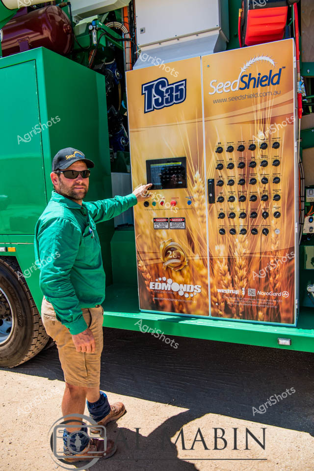 A man working as a farm worker operates controls on a SeedShield seed cleaning unit mounted on a green truck during harvest operations in Western Australia. The bright orange equipment panel displays numerous selection buttons and controls for wheat processing, while the worker in green clothing and cap demonstrates the precision technology used in modern cropping systems. This close-up view captures the technical sophistication of seed cleaning equipment essential for grain quality management in WA's wheatbelt region.