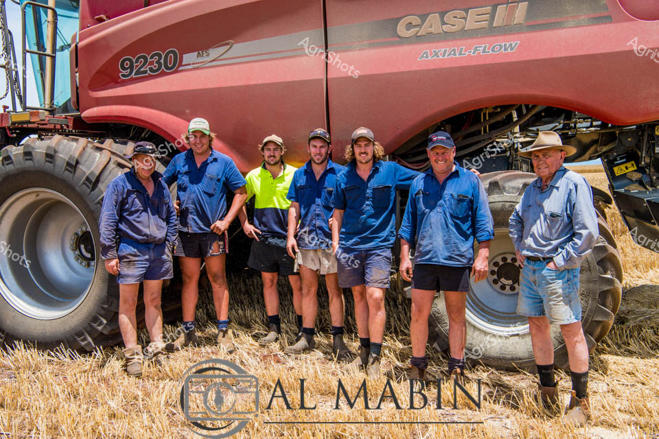 A group of seven farm workers stands proudly beside a massive red Case IH 9230 Axial-Flow combine harvester in a Western Australian wheat field. The men, dressed in blue work shirts and shorts typical of Australian harvest crews, pose together on the stubbled ground after completing their grain harvesting operations. The enormous header, with its distinctive red bodywork and large flotation tires, dominates the scene and represents the industrial scale of modern wheat production in WA's wheatbelt region. Golden wheat stubble carpets the surrounding paddock, indicating a successful harvest season where this powerful machinery has efficiently gathered the grain crop.