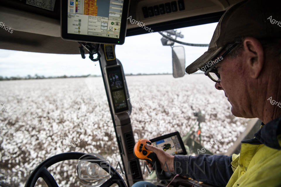 Cotton Harvest: Precision Agriculture in Queensland

A weathered agricultural worker sits in the climate-controlled cab of a sophisticated John Deere cotton harvester, surrounded by a pristine white sea of cotton bolls stretching across the Queensland landscape. Dressed in a yellow safety jacket and cap, the operator navigates the massive machine with practiced expertise, using multiple digital displays and a handheld control device to monitor harvesting performance. The interior of the harvester is a testament to modern agricultural technology, with electronic screens displaying complex operational data and GPS tracking systems guiding each precise pass through the cotton field. Brilliant white cotton fields extend to the horizon, creating a stark contrast against the soft, muted tones of the distant landscape. The operator's focused posture and careful interaction with the machine's sophisticated controls reveal the high-tech skill required in contemporary cotton production, where precision, efficiency, and technological mastery are as critical as traditional farming knowledge.