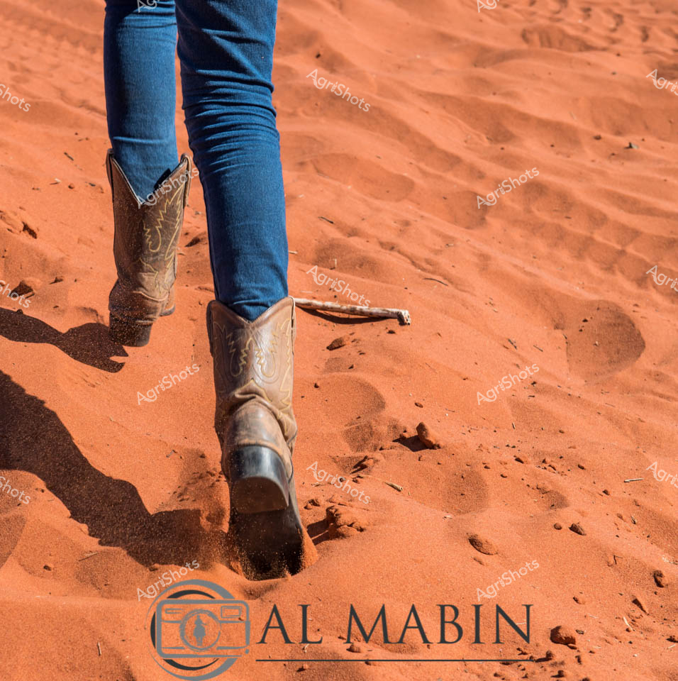 Weathered leather boots sink into the rich, rust-colored sands of the Australian outback, casting long shadows across the undulating desert terrain. The meticulously crafted cowboy boots, adorned with intricate western-style stitching, tell a story of resilience and rugged agricultural life. Slim-fit blue denim jeans contrast against the vibrant ochre landscape, suggesting the enduring spirit of station workers who navigate challenging environments with strength and purpose. Each footprint leaves a momentary mark on the shifting desert floor, a testament to the constant movement and hard work of rural professionals. This image embodies the raw beauty of rural Australia - a landscape of extreme conditions and unwavering human determination.