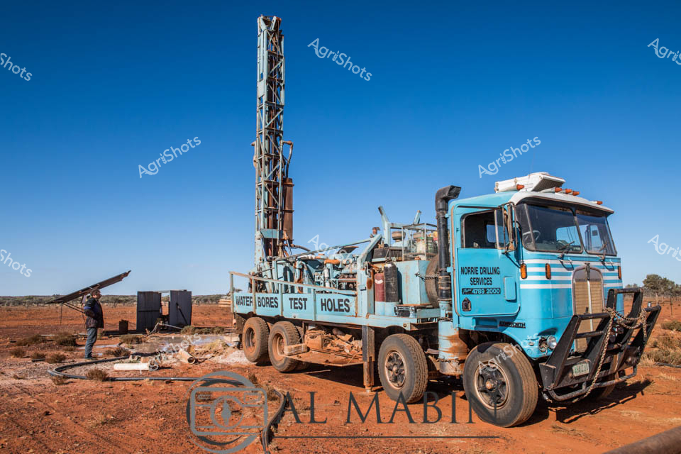 Beneath an electric blue outback sky, a robust Norrie Drilling Services water bore truck stands resolute on the rust-red earth of remote Western Australia. The light blue truck, mounted with an imposing telescopic drilling rig, represents critical infrastructure for rural water exploration, strategically positioned in an arid landscape where groundwater access is lifeline for agricultural survival. A worker in dark protective clothing stands near the vehicle, overseeing the delicate process of drilling test holes that will potentially unlock underground water resources for pastoral stations and agricultural enterprises. The heavy-duty truck, with its rugged front bull bar and industrial drilling apparatus, embodies the technical precision and resilience required to sustain agriculture in challenging desert environments. Stark white lettering on the vehicle's azure body declares its specialized service, while the surrounding terrain - scattered with low scrub and rocky ground - speaks to the unforgiving nature of Australian inland territories. This scene captures the essential technical intervention that enables human productivity in some of the continent's most demanding agricultural frontiers.
