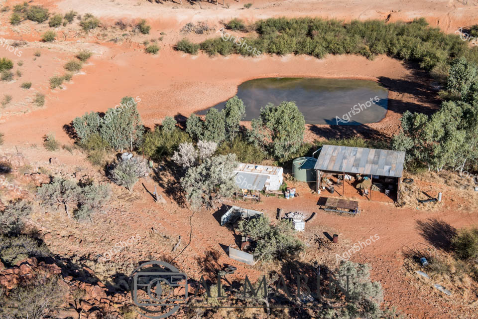 Arid Outback Water Hole and Homestead

Rusty red earth cradles a precious water hole surrounded by silvery-green mulga and saltbush, capturing the resilient heart of a remote Australian pastoral station. Weathered tin sheds and water tanks cluster beside the dark, life-giving dam, their sun-bleached surfaces telling stories of generations of bush survival. The aerial perspective reveals the stark beauty of inland Australia - a landscape of muted ochre and sage, where every drop of water means survival for livestock and livelihood. Rugged infrastructure speaks to the practical engineering of station life, with carefully positioned buildings offering shelter and strategic water access in this unforgiving terrain. Shadows stretch long across the rocky ground, suggesting early morning or late afternoon light that softens the harsh desert environment. This is a quintessential snapshot of outback agricultural persistence - a testament to human adaptation and agricultural resilience in one of the world's most challenging farming landscapes.