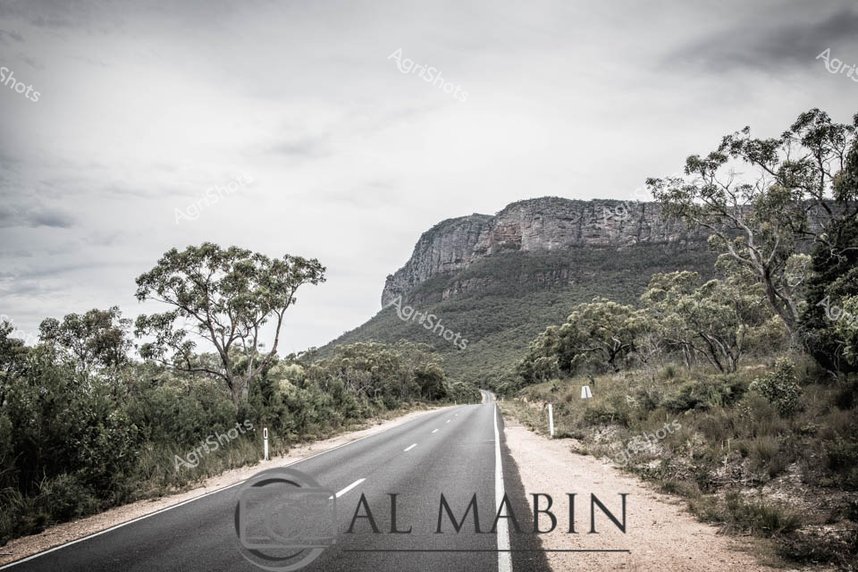 Empty rural highway cutting through dense eucalyptus woodland, leading towards an imposing rocky escarpment under a pale, overcast sky. The sealed bitumen road winds between scattered gum trees and low scrubland, creating a stark contrast between the smooth black asphalt and the rugged native landscape. Weathered road markers line the verge, guiding travelers through this remote Victorian bushland setting. Silvery-green foliage and granite cliff faces speak to the harsh beauty of Australia's interior regions, capturing the essence of rural connectivity and isolation. The muted color palette and dramatic geological backdrop evoke a sense of quiet anticipation, as if the road itself is a narrative waiting to unfold across the expansive landscape. This image epitomizes the raw, untamed character of regional Victoria, where infrastructure meets wilderness in a timeless Australian tableau.