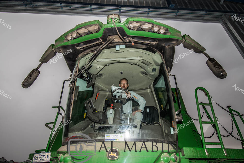 A person sits in the cab of a green John Deere header during grain harvest operations in Victoria. The harvester's cab is positioned prominently in the frame, showing the modern interior and operator controls of this essential cropping equipment. The header represents the backbone of Australia's broadacre grain industry, designed for efficient harvesting of wheat, barley, and other cereal crops across the Victorian farming regions.