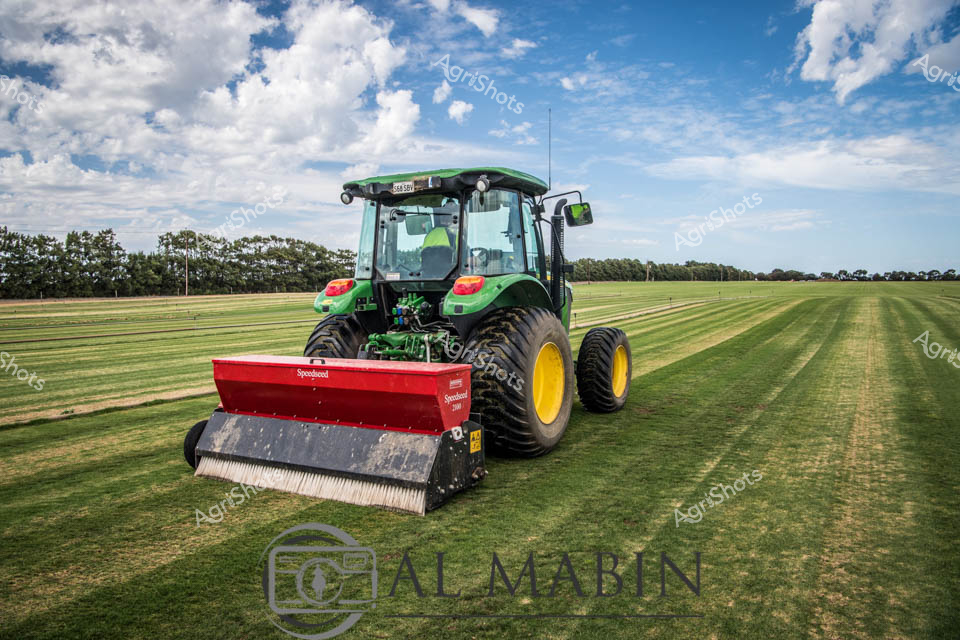 Emerald-green John Deere tractor glides across a meticulously maintained turf field under a billowing cloudscape of azure and white. The robust agricultural machine carries a vibrant red Redexim Speedseed implement, its brushed attachment creating precise stripes across the verdant grass surface. Golden yellow wheels provide striking contrast against the lush green landscape, suggesting professional groundskeeping or sports field maintenance in the expansive South Australian countryside.

Crisp morning light illuminates the scene, casting soft shadows and highlighting the geometric precision of the machine's path. The background reveals a serene rural setting with distant tree lines marking the horizon, creating a sense of agricultural mastery and careful land management. The tractor's advanced design and specialized seeding equipment showcase modern agricultural technology's sophistication, demonstrating how precision and innovation transform landscape maintenance.

The immaculate field and powerful machinery communicate a narrative of meticulous care, technological advancement, and the skilled craft of agricultural professionals who transform natural landscapes with expertise and purpose.