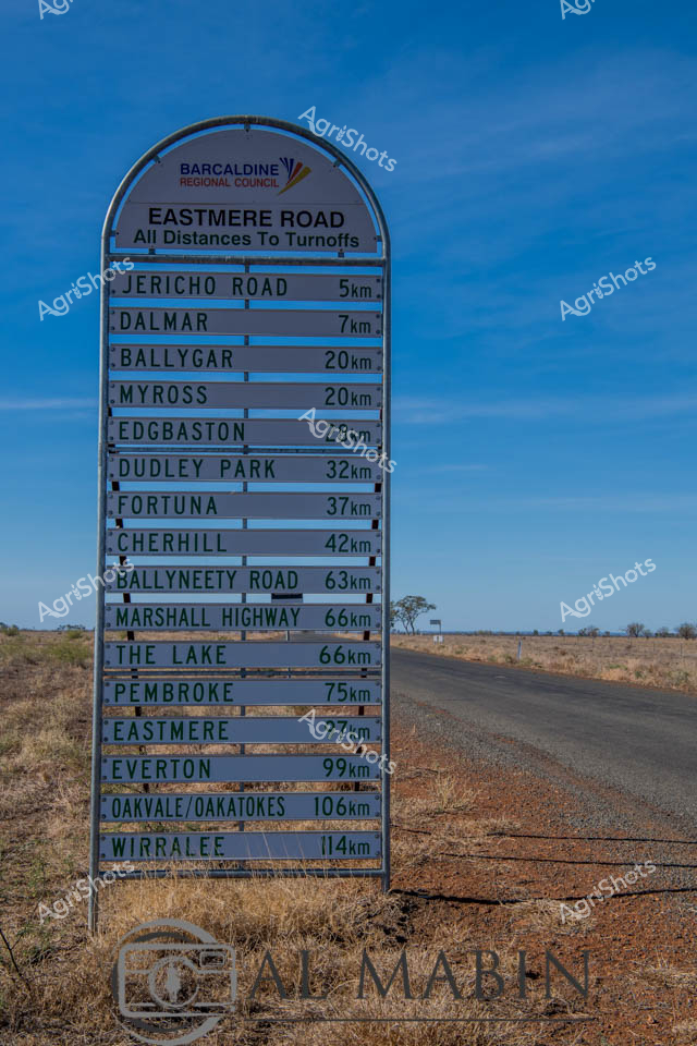 A weathered distance sign stands sentinel on the Eastmere Road, marking the vast distances across the arid Queensland landscape. The outback Queensland Regional Council signpost chronicles a remarkable journey through the sparse, golden-brown rural terrain, with distances stretching from the nearby Jericho Road at 5km to Wirralee at 114km. Rugged grasslands and sparse vegetation frame the unsealed road, which cuts a straight line through the expansive dry pastoral country, capturing the essence of remote Australian agricultural regions.

The blue-backed sign, bleached by relentless sunlight, lists a series of rural localities that map the complex network of stations and settlements across this challenging landscape. Distant destinations like Ballygar, Myross, and Dudley Park punctuate the sign, each kilometer representing the immense scale of agricultural enterprise in central Queensland. A lone tree stands in the background, a subtle testament to the resilience required to survive in this harsh but beautiful environment.

This image epitomizes the spirit of the Australian outback - vast, uncompromising, and fundamentally tied to the agricultural rhythms that define rural life. The sign serves not just as a navigational marker, but as a cultural artifact documenting the interconnected communities of the region.