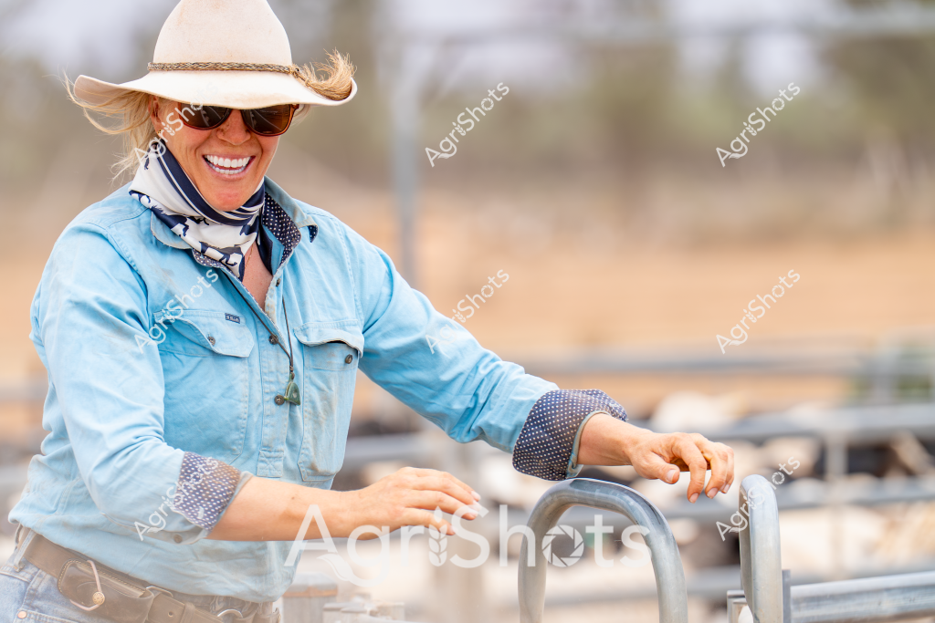 A woman farmer in a light blue chambray shirt and white wide-brimmed hat stands at a metal gate in an arid rural landscape. Her sunglasses and cheerful smile suggest confidence and enjoyment while working outdoors, with a blurred background of dry paddocks and scattered trees. She is wearing practical workwear with rolled chambray sleeves and a patterned navy scarf, typical of Australian agricultural attire. The soft focus and warm tones indicate this is likely a late morning or early afternoon scene during a dry season. Her relaxed but alert posture and positioning near rural infrastructure conveys her connection to the land and her professional role in managing the property.