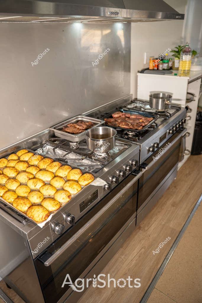 A professional kitchen stove is loaded with multiple cooking stages: golden-brown roasted potatoes occupy one section, perfectly aligned in neat rows, while two trays of seared meat are cooking on the adjacent burners. Stainless steel pots simmer alongside the dishes, suggesting a precise and methodical cooking process. The commercial-grade stove features multiple gas burners and a gleaming stainless steel backsplash, creating a clean and professional cooking environment. Cooking oils and condiment bottles are visible in the background, indicating a well-equipped kitchen preparation area. The wooden floor and sleek appliances suggest a modern, efficient kitchen setup designed for high-quality food preparation.

The image captures the precision and organization of professional cooking, with each element carefully positioned and cooked to perfection. The golden potatoes - likely roasted with a crisp exterior and fluffy interior - contrast beautifully with the rich, seared meat, showcasing the technical skill and attention to detail required in professional culinary work.