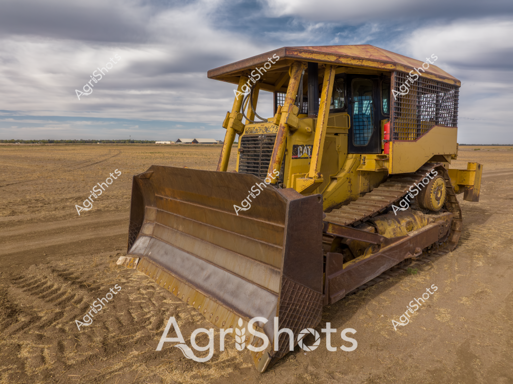 A yellow Caterpillar bulldozer sits positioned on a dry, expansive agricultural landscape under a cloudy, dramatic sky. The heavy-duty construction equipment, painted in its distinctive bright yellow color, features protective metal mesh guards on the operator's cabin and a large, sturdy blade for earthmoving and land preparation. Mounted on robust tracked wheels, the bulldozer appears ready for agricultural or construction work, with its blade leaving distinct tread marks in the dusty, light brown soil. The surrounding landscape is a broad, flat terrain with distant farm structures visible on the horizon, suggesting this is likely a rural farming or agricultural development site. The machine's weathered appearance - with some rust and wear on the blade and body - indicates it has seen significant use in challenging outdoor conditions. The overcast sky with layers of gray and white clouds adds a moody atmospheric backdrop to this powerful piece of agricultural and construction equipment.