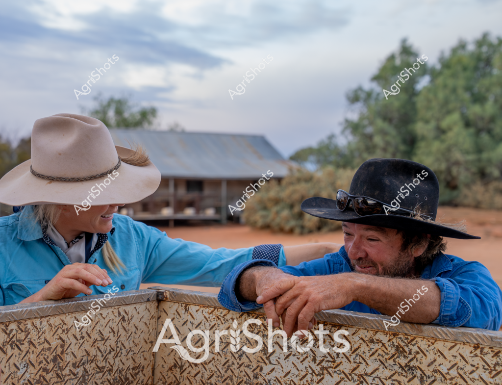 A man and a woman in blue work shirts and wide-brimmed hats lean against a textured metal container in a rural Australian setting. The scene captures a moment of quiet conversation on what appears to be a remote station, with a rustic shed and trees visible in the background. The woman wears a light-colored wide-brimmed hat, while the man sports a black hat with sunglasses, both dressed in practical workwear typical of Australian rural life. The reddish earth and relaxed posture suggest a pause during a long day of farm work, with the metal container providing a focal point for their interaction. The overcast sky and surrounding landscape evoke the vast, understated beauty of the Australian outback, highlighting the connection between rural workers and their challenging environment.