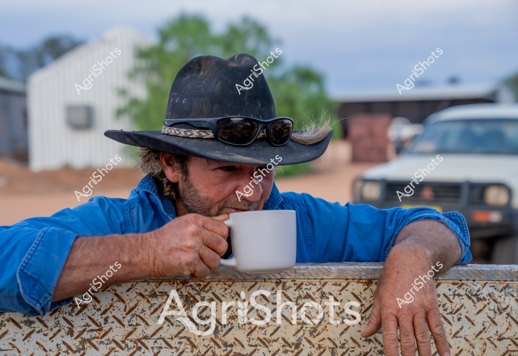 A man stockman takes a contemplative coffee break, leaning against a textured metal surface in a rural farm setting. Wearing a classic black cowboy hat and sunglasses, he's dressed in a vivid blue work shirt that suggests a day of agricultural labor. The background reveals farm infrastructure - a white building, parked vehicle, and scattered trees - typical of an Australian rural property. His posture - one hand gripping a white coffee mug, the other resting casually - conveys a moment of quiet reflection during a busy workday. The rugged, weathered aesthetic speaks to the resilience and character of Australian farm workers, capturing a slice of everyday rural life with an authentic, unposed feel.