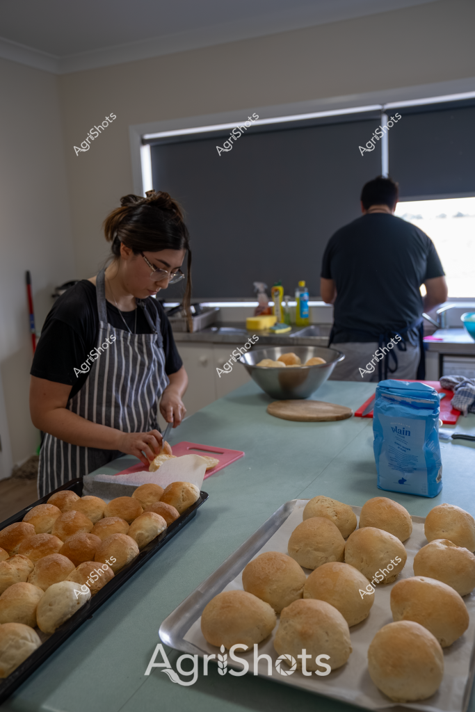 A woman in a striped apron and glasses carefully prepares bread rolls in a home kitchen, working methodically alongside another person. Multiple trays of golden-brown bread rolls are arranged across the kitchen bench, suggesting a baking session in progress. A blue bag of flour sits nearby, indicating fresh ingredients are being used. The kitchen has a modern, clean appearance with neutral walls and a roller blind, creating a calm and organized workspace. The scene captures a moment of culinary preparation, with precise movements and careful attention to detail evident in the baker's work.

The bread rolls appear uniformly shaped and have a rich, even coloration, suggesting skilled baking technique. The kitchen environment seems purposeful and focused, with two people working together in a coordinated manner. A bowl of additional ingredients is visible, and cooking oils are positioned in the background, completing the mise en place for what looks like a bread-making project.
