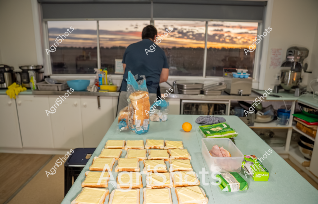 A man in a navy blue shirt prepares sandwiches in a professional kitchen, with a stunning sunset visible through large windows behind him. Slices of white bread are meticulously arranged on a pale blue work surface, alongside ingredients including a container of ham, an orange, and green packaged items. Commercial kitchen equipment like mixers and stainless steel preparation surfaces are visible in the background, suggesting this is a food preparation area with professional-grade facilities. The warm evening light streams through the windows, casting a golden glow across the workspace and creating a serene backdrop for the sandwich-making process. The organized layout and careful arrangement of ingredients highlight the precision and methodical approach to food preparation in this commercial kitchen setting.