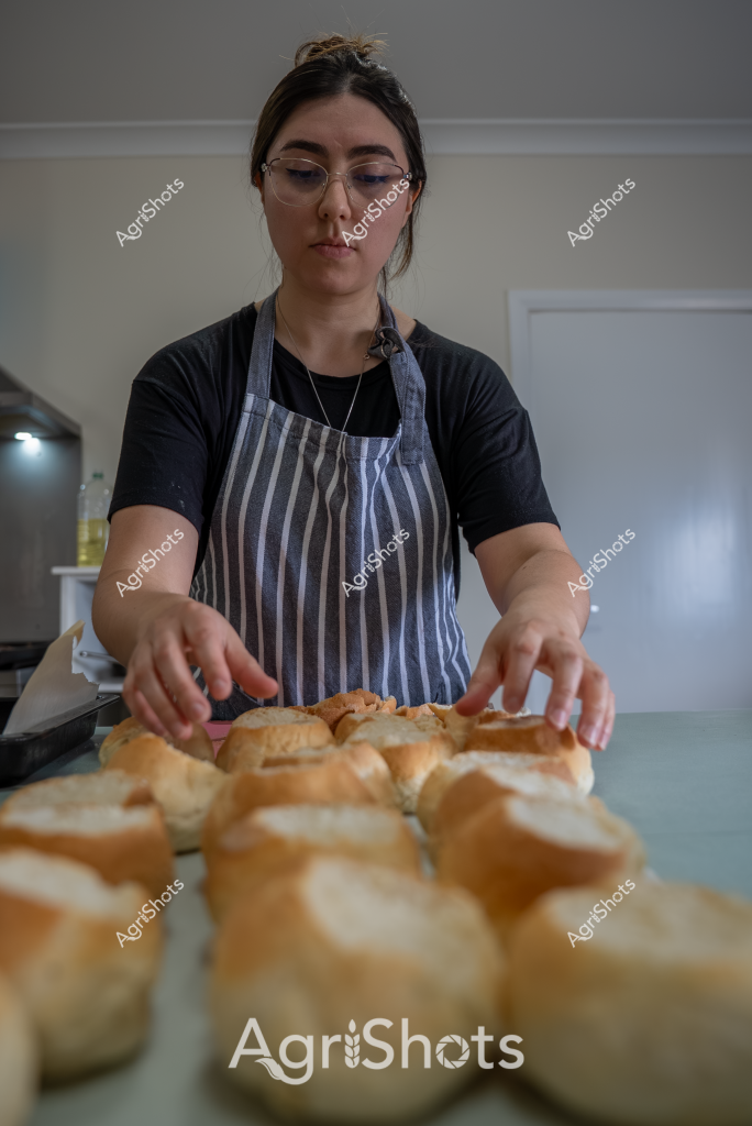 A woman baker carefully arranges freshly baked bread rolls on a kitchen counter, wearing a striped gray apron over a black shirt and glasses. Her focused expression and precise hand movements suggest professional skill and attention to detail as she positions the golden-brown rolls with care. The bread appears to be artisan-style white rolls with a crisp, slightly dusted crust, reflecting traditional baking techniques. The kitchen environment is clean and neutral, with soft lighting highlighting the warm tones of the fresh bread. Her positioning suggests she is preparing these rolls for sale, potentially for a bakery, café, or local market. The image captures a moment of culinary craftsmanship, showcasing the precision and dedication involved in artisan bread preparation.