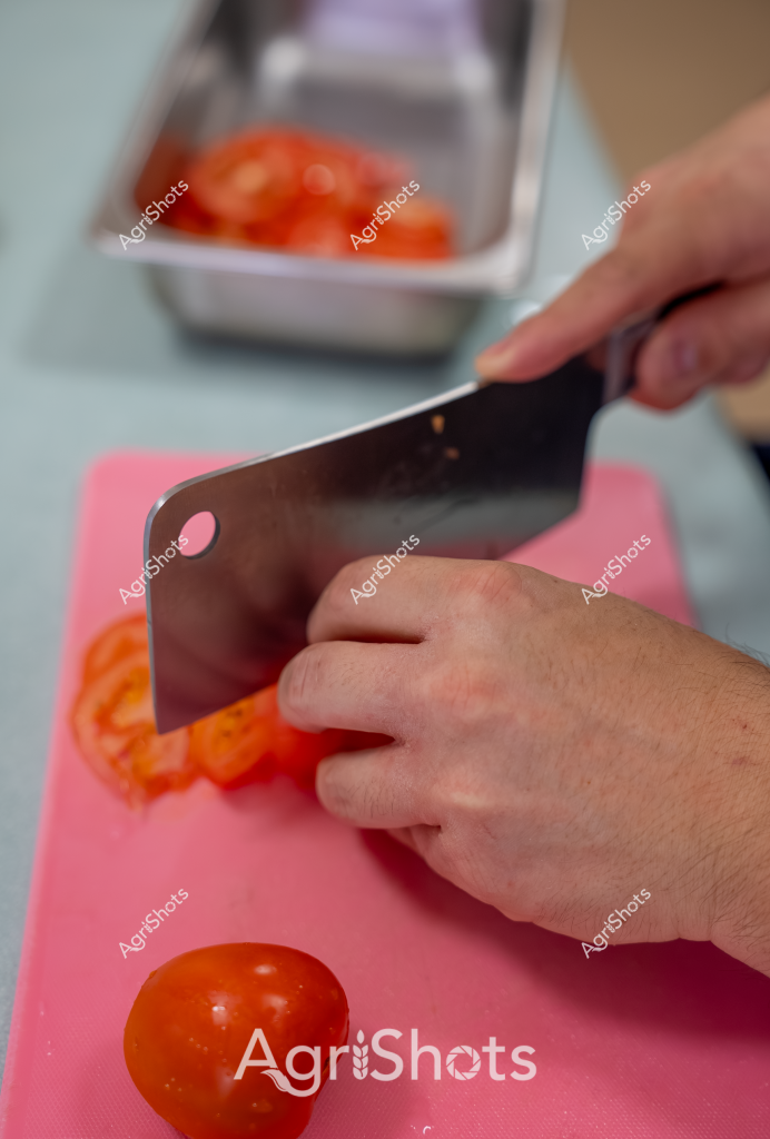 A hand uses a sharp kitchen knife to slice ripe, bright red tomatoes on a pink cutting board. In the background, a metal tray contains additional whole tomatoes, creating a vibrant culinary scene focused on fresh produce preparation. The close-up shot captures the precise moment of slicing, with the gleaming knife blade positioned to cut through the juicy tomato. The bright colors - the vivid red of the tomatoes and the pink cutting surface - create a visually appealing composition that suggests fresh food preparation in a home or professional kitchen setting. The carefully positioned hand demonstrates careful, controlled cutting technique, highlighting the attention to detail in food preparation.