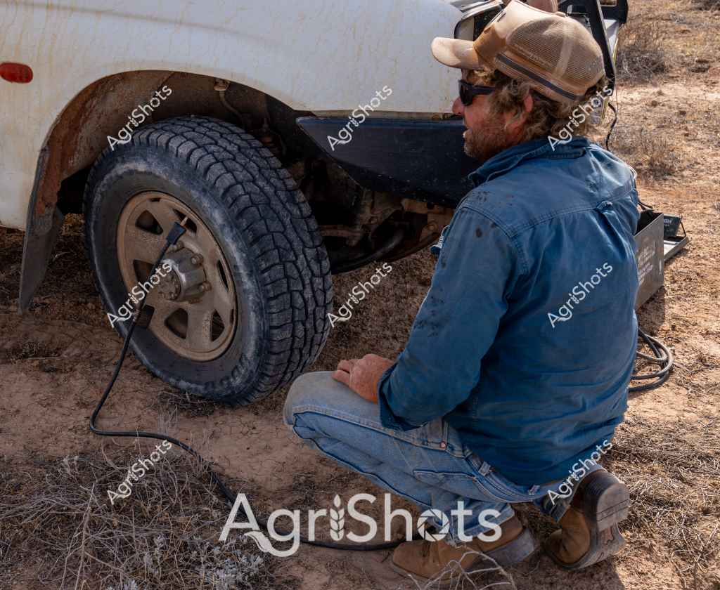 A man wearing a blue work jacket and tan cap carefully examines a vehicle tire while crouched on dry, rocky ground. The white vehicle has a rusted wheel arch, and the tire appears to be an all-terrain model with deep tread patterns suitable for rough rural terrain. He is wearing sunglasses and work boots, suggesting he is performing roadside maintenance or vehicle inspection in an isolated, arid landscape. The surrounding environment looks like a typical Australian outback or remote farming region, with sparse vegetation and dusty ground indicating challenging environmental conditions. The man's posture and attentive positioning suggest he is experienced in dealing with vehicle repairs in challenging outdoor settings.