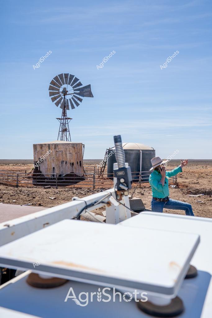 A woman in a teal shirt and wide-brimmed hat stands near a classic windmill and water tank in a stark, arid landscape. The windmill's metal blades rise against a clear blue sky, positioned next to a weathered cylindrical water tank enclosed by a metal fence. In the foreground, scientific or surveying equipment is visible, suggesting the woman is conducting environmental or agricultural research in this remote, dry terrain. The landscape appears to be a typical Australian outback scene - flat, dusty ground stretching to a distant horizon, with minimal vegetation indicating challenging drought conditions. The bright turquoise of the woman's shirt provides a vivid contrast to the muted browns and grays of the surrounding environment, highlighting human presence in this harsh but essential agricultural setting.